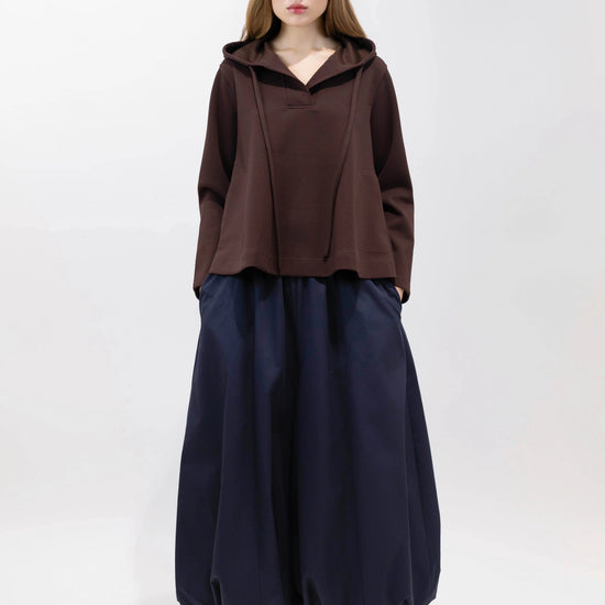 A model stands against a plain white studio background wearing a dark brown cropped hooded top, loose dark navy wide-leg pants, and brown sneakers.