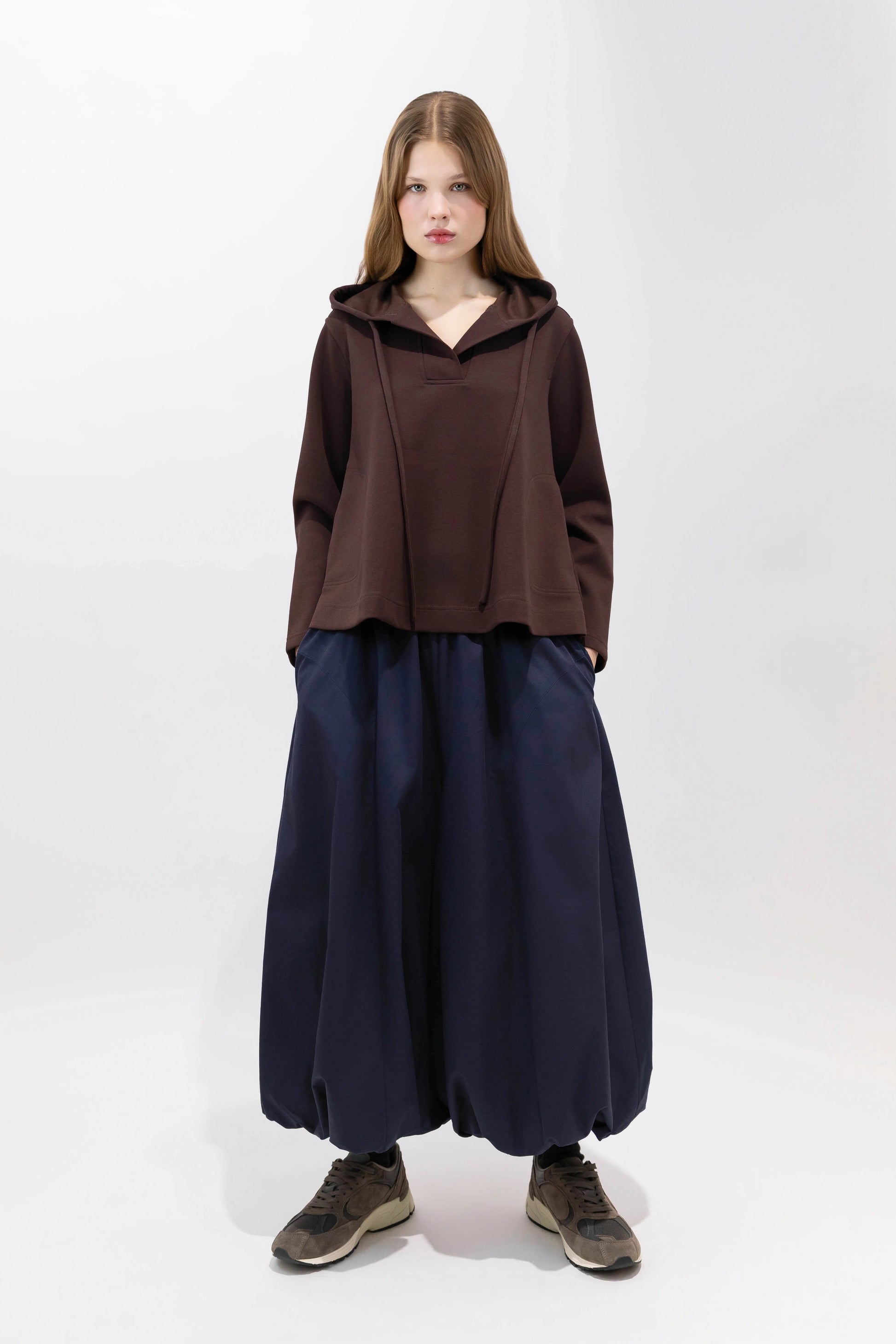 A model stands against a plain white studio background wearing a dark brown cropped hooded top, loose dark navy wide-leg pants, and brown sneakers.