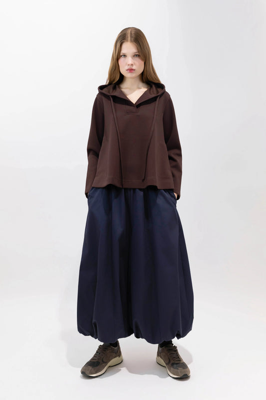 A model stands against a plain white studio background wearing a dark brown cropped hooded top, loose dark navy wide-leg pants, and brown sneakers.