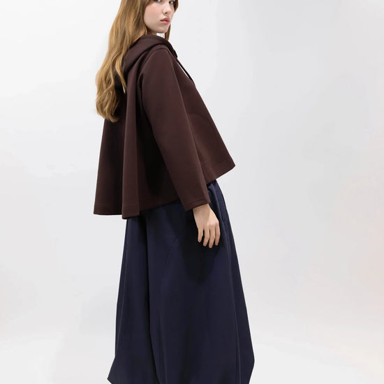 A model is shown from the back wearing a dark brown cape-style jacket, a long navy blue skirt, and brown sneakers against a plain white studio background.