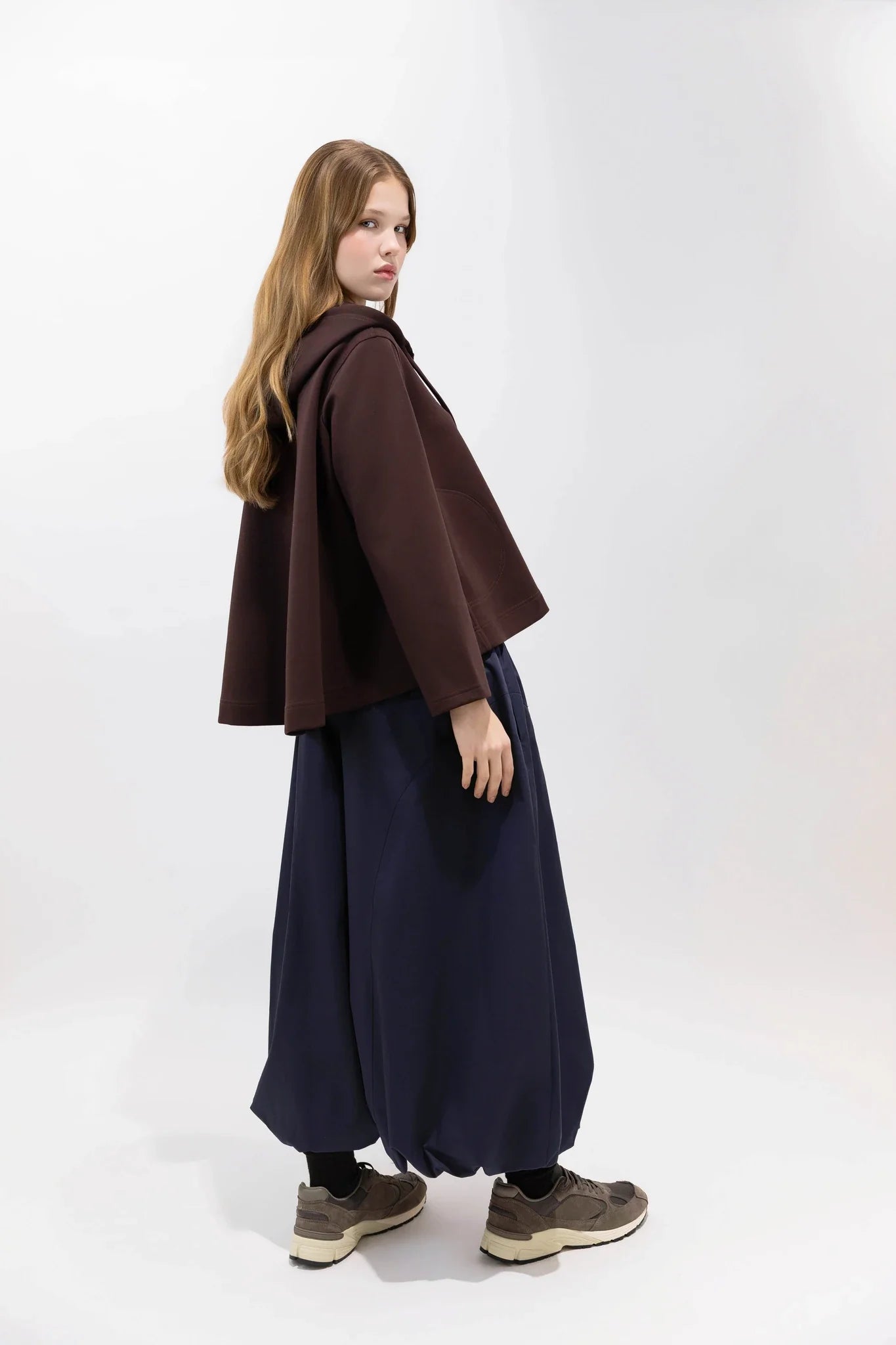 A model is shown from the back wearing a dark brown cape-style jacket, a long navy blue skirt, and brown sneakers against a plain white studio background.