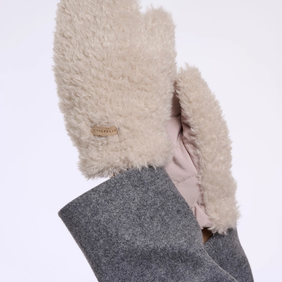 A close-up of a person's hand wearing fluffy cream-colored shearling mittens with a gray wool-lined cuff, set against a plain white background.