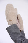 A close-up of a person's hand wearing fluffy cream-colored shearling mittens with a gray wool-lined cuff, set against a plain white background.