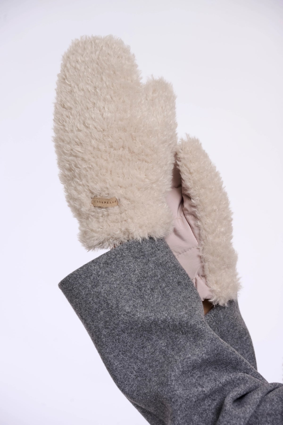 A close-up of a person's hand wearing fluffy cream-colored shearling mittens with a gray wool-lined cuff, set against a plain white background.