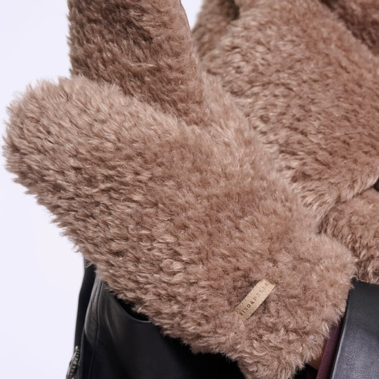 A close-up of a person wearing fluffy taupe-brown faux-fur gloves over a black leather jacket and matching brown coat, with a small fabric brand tag visible on one cuff.