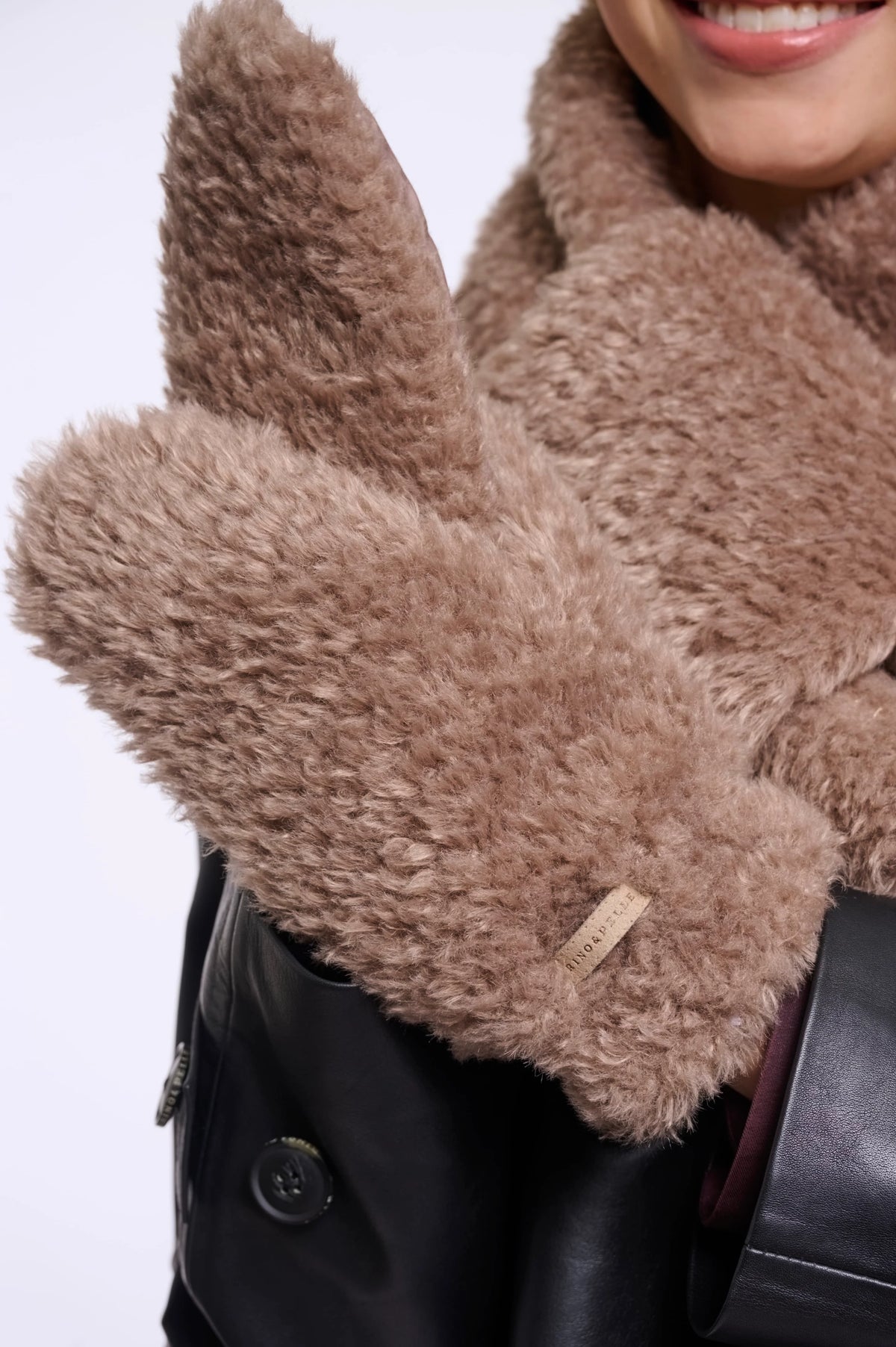 A close-up of a person wearing fluffy taupe-brown faux-fur gloves over a black leather jacket and matching brown coat, with a small fabric brand tag visible on one cuff.