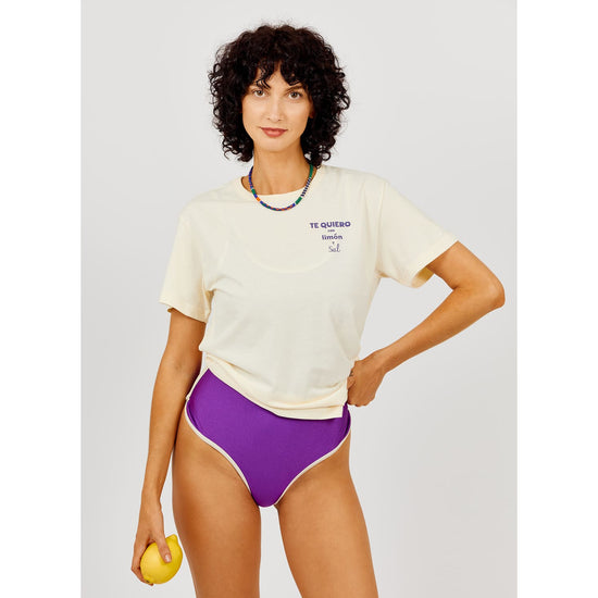 Woman wearing a white t-shirt with text and purple high-waisted bikini bottoms, holding a yellow object.