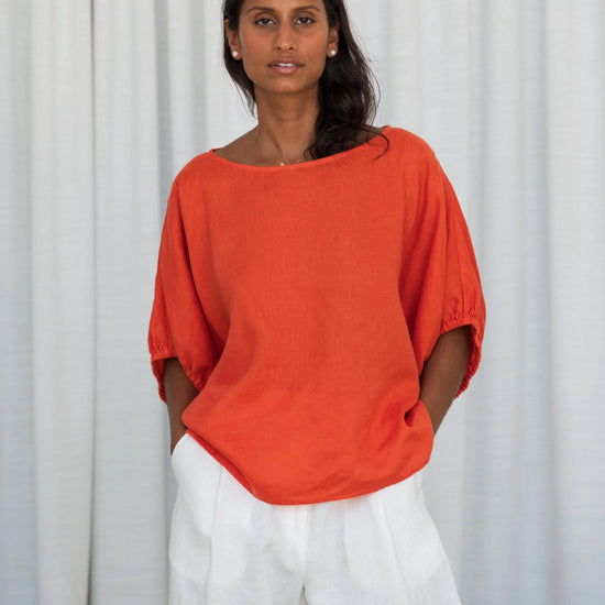 Woman wearing an orange top and white pants against a white curtain background from Lilly Pilly