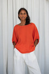 Woman wearing an orange top and white pants against a white curtain background from Lilly Pilly