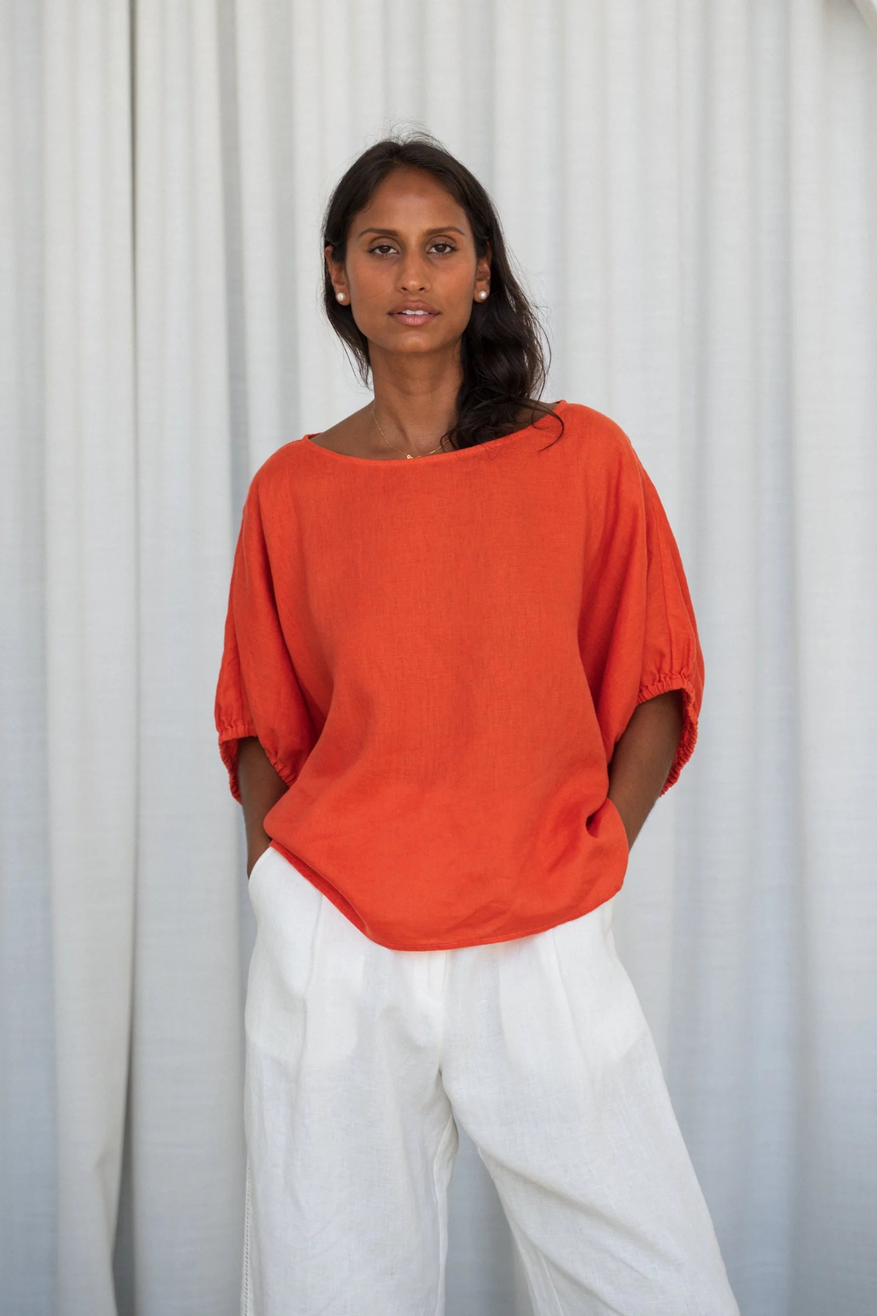 Woman wearing an orange top and white pants against a white curtain background from Lilly Pilly