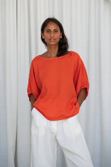 Woman wearing an orange top and white pants against a white curtain background from Lilly Pilly