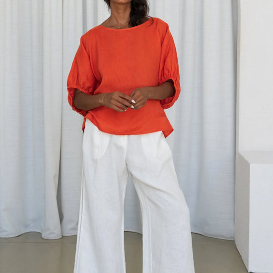 Woman wearing an orange top and white pants standing against a white curtain from Lilly Pilly