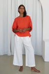 Woman wearing an orange top and white pants standing against a white curtain from Lilly Pilly