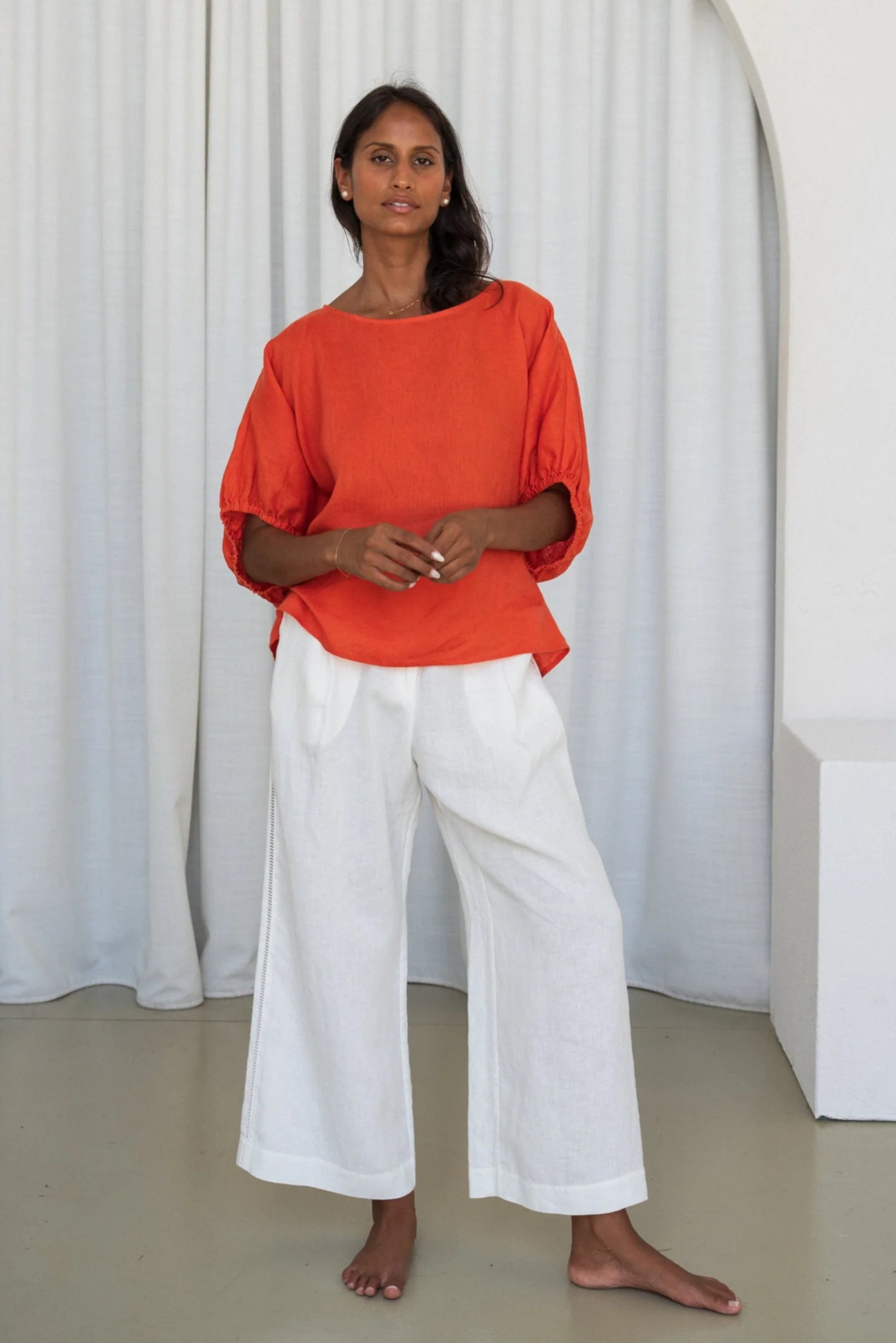 Woman wearing an orange top and white pants standing against a white curtain from Lilly Pilly