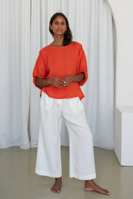 Woman wearing an orange top and white pants standing against a white curtain from Lilly Pilly
