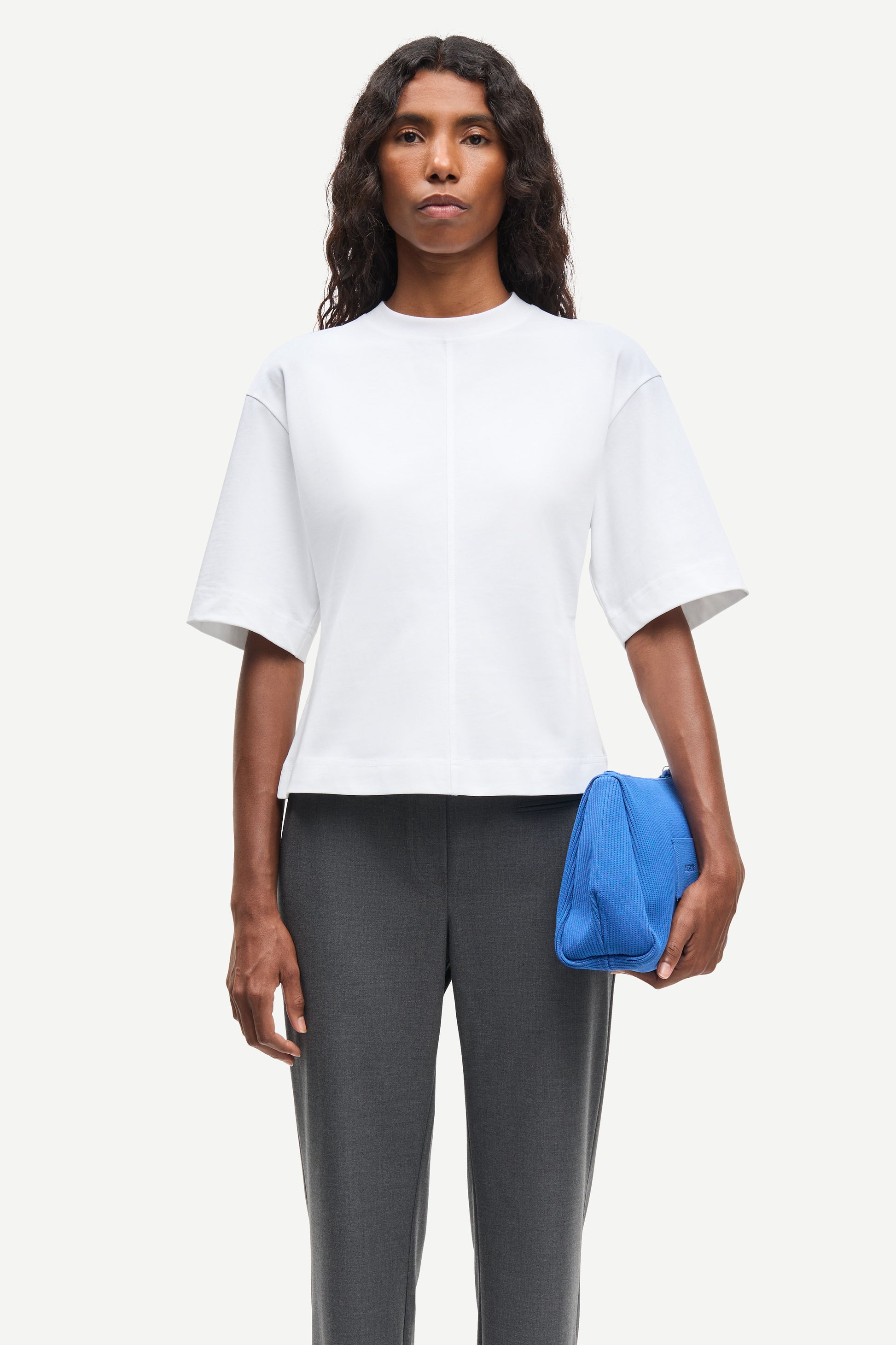 Woman wearing a white top and gray pants, holding a blue clutch against a white background from Samsoe Samsoe