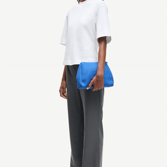 Woman wearing a white top and gray pants holding a blue clutch against a white background from Samsoe Samsoe
