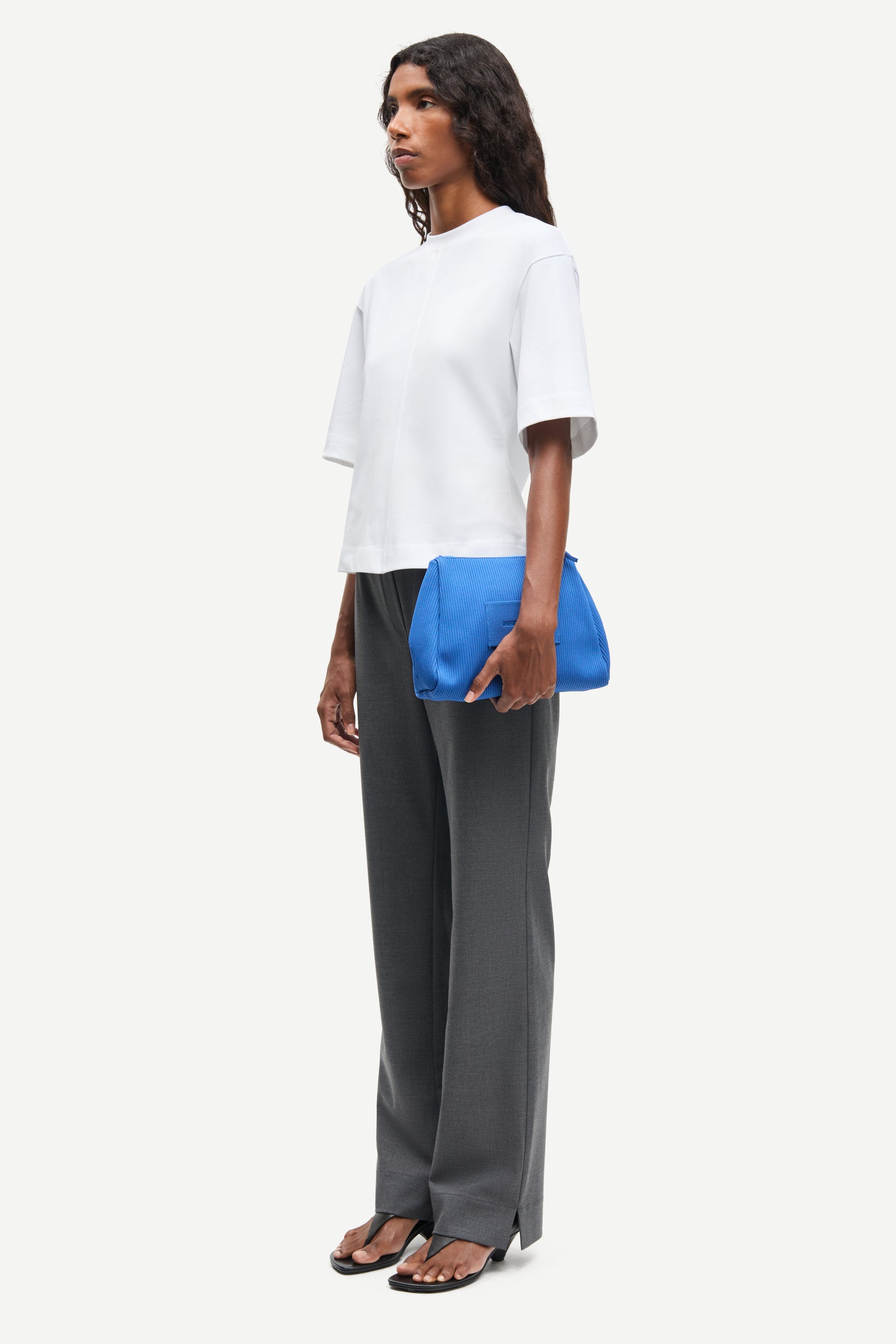 Woman wearing a white top and gray pants holding a blue clutch against a white background from Samsoe Samsoe