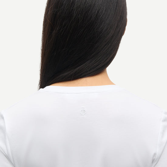 Person with long black hair from behind on a light gray background wearing white samsoe samsoe round neck long sleeve tee