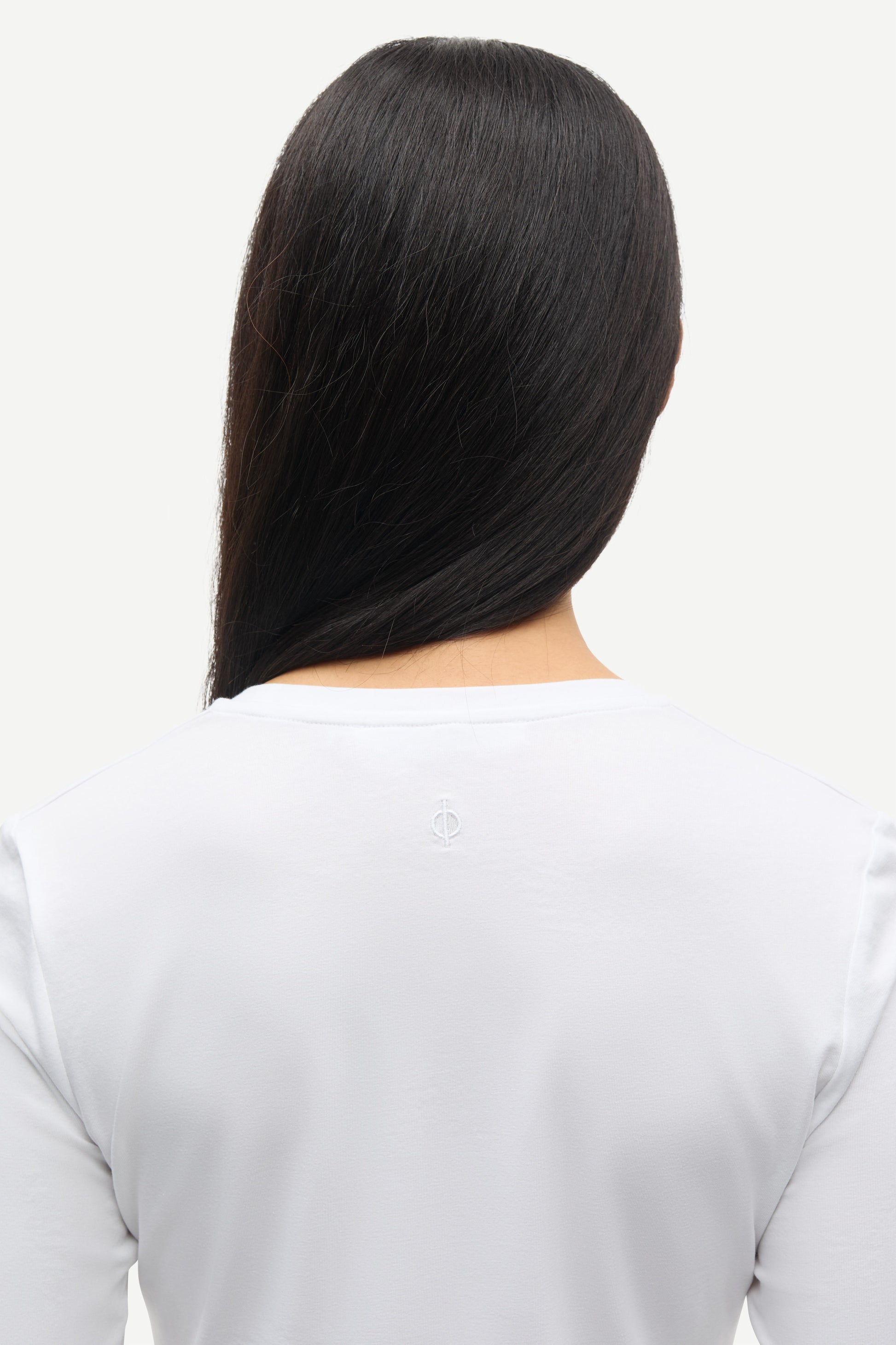 Person with long black hair from behind on a light gray background wearing white samsoe samsoe round neck long sleeve tee