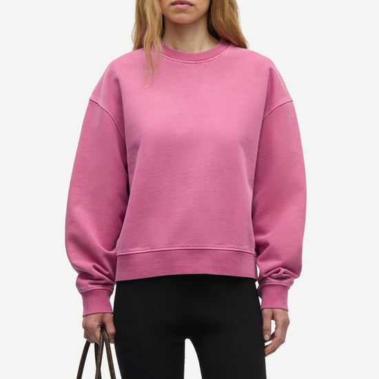 A woman modeling a dusty pink crewneck sweatshirt with dropped shoulders and ribbed cuffs, paired with black leggings and holding a dark brown leather tote bag against a light gray studio background.