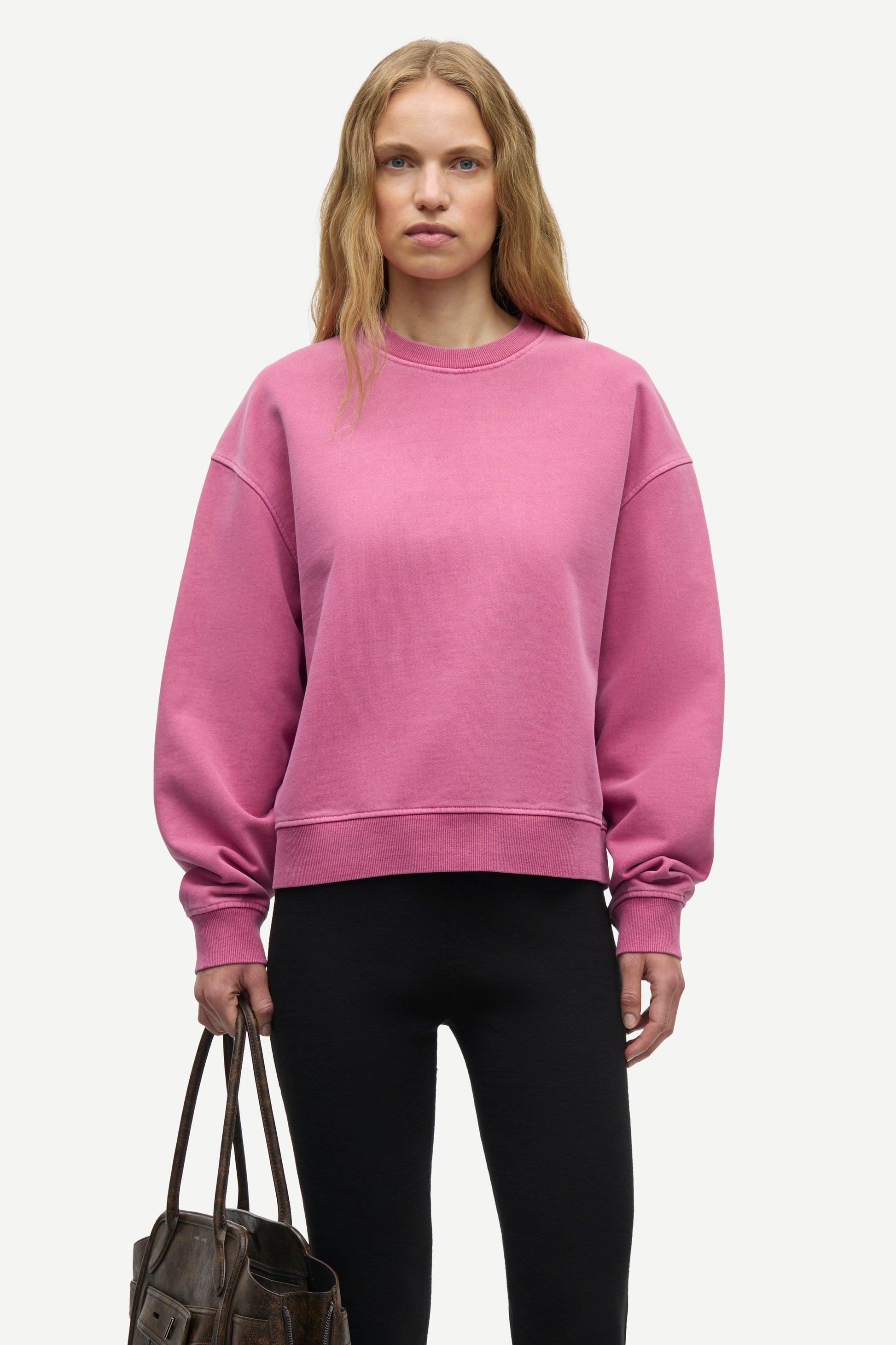 A woman modeling a dusty pink crewneck sweatshirt with dropped shoulders and ribbed cuffs, paired with black leggings and holding a dark brown leather tote bag against a light gray studio background.