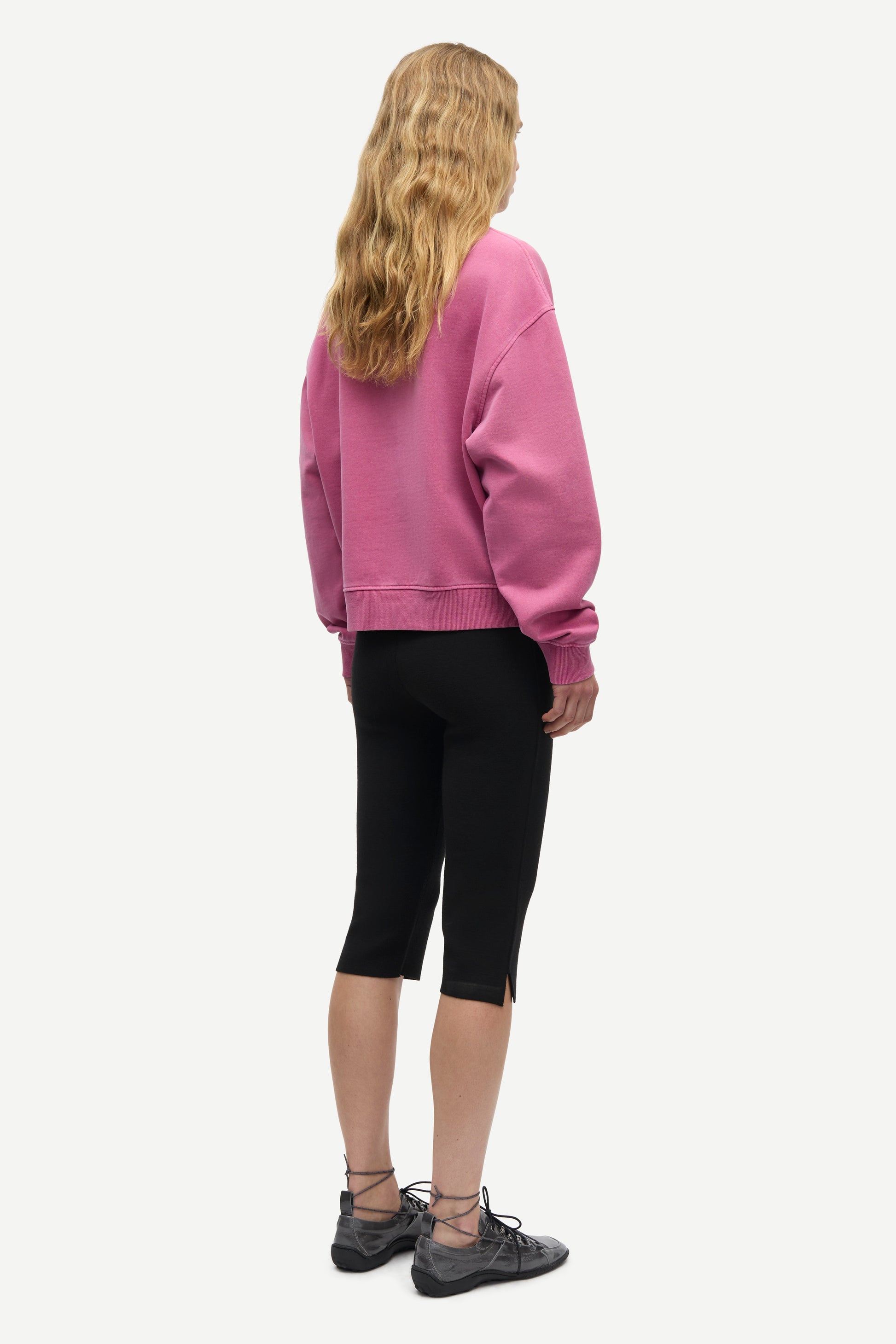 A woman shown from the back wearing a dusty pink crewneck sweatshirt, black capri leggings, and dark gray lace-up shoes against a light gray background.