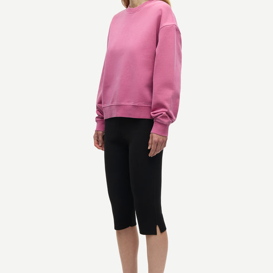 A woman modeling a dusty pink crewneck sweatshirt, black cropped capri pants, and dark gray lace-up sneakers against a plain light gray background.