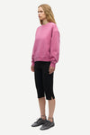 A woman modeling a dusty pink crewneck sweatshirt, black cropped capri pants, and dark gray lace-up sneakers against a plain light gray background.