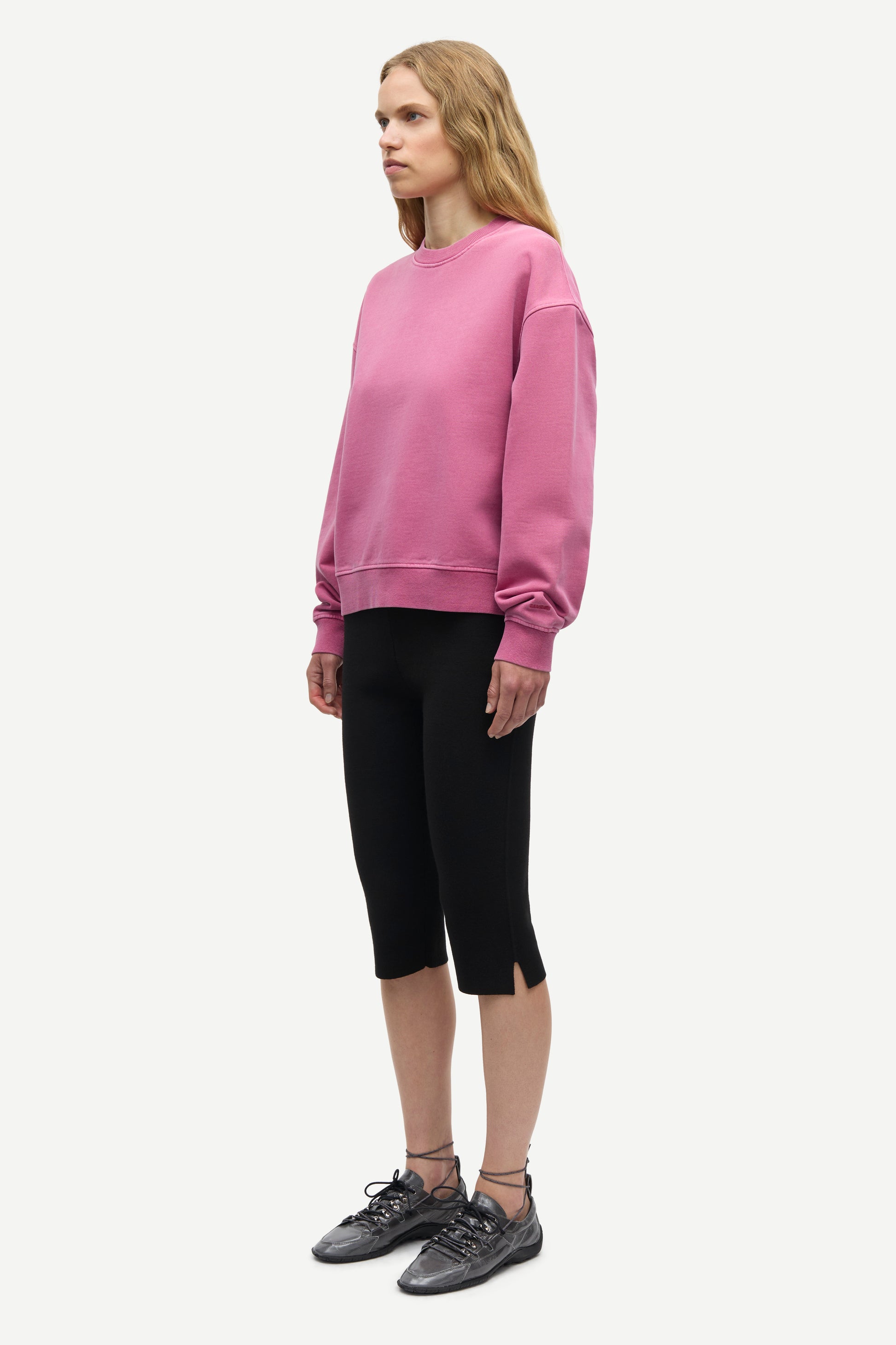 A woman modeling a dusty pink crewneck sweatshirt, black cropped capri pants, and dark gray lace-up sneakers against a plain light gray background.