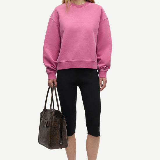 A model stands against a light gray studio background wearing a dusty rose pink crewneck sweatshirt, black cropped leggings, and black sneakers, while holding a dark brown leather tote bag.