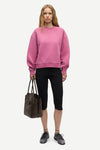 A model stands against a light gray studio background wearing a dusty rose pink crewneck sweatshirt, black cropped leggings, and black sneakers, while holding a dark brown leather tote bag.