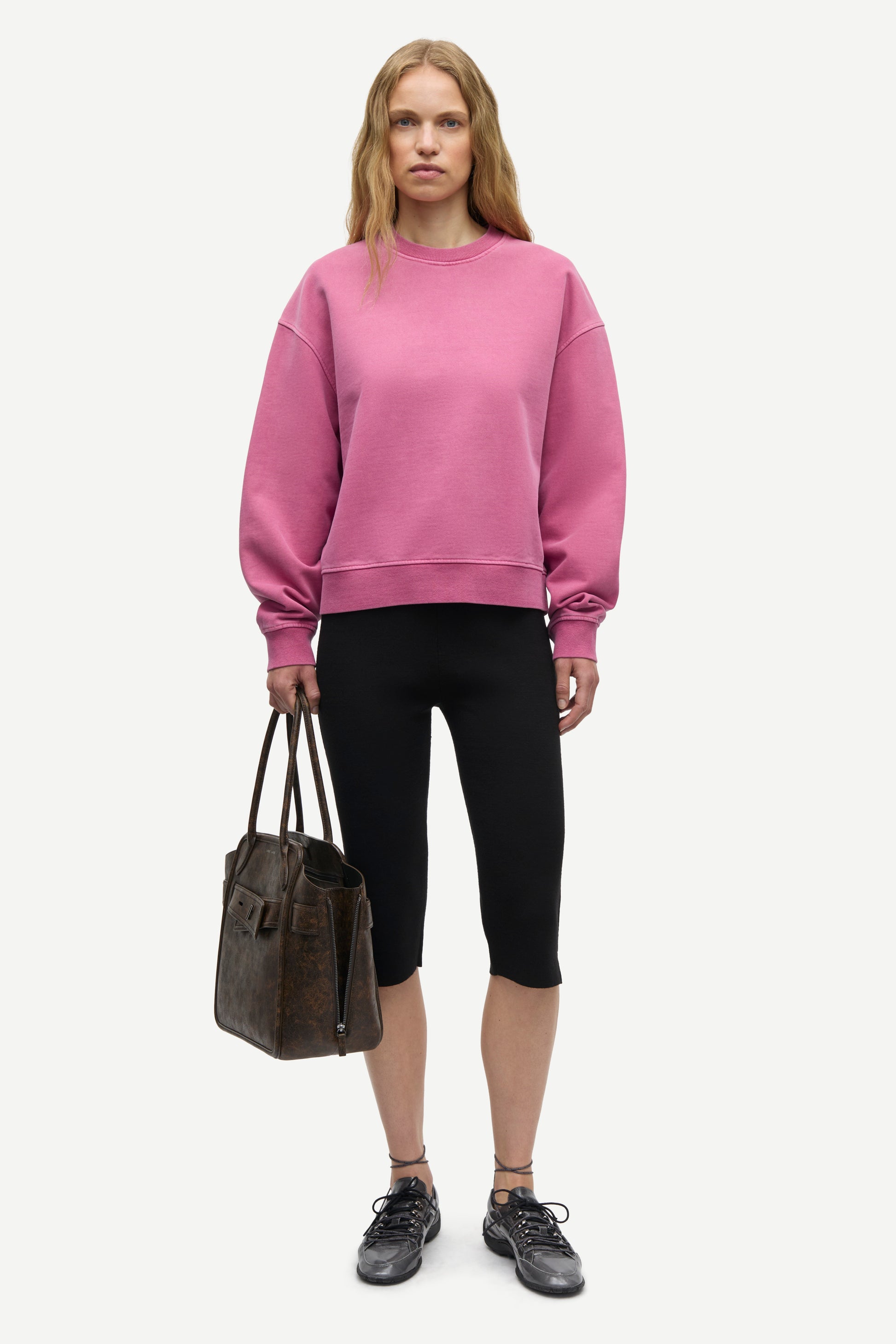 A model stands against a light gray studio background wearing a dusty rose pink crewneck sweatshirt, black cropped leggings, and black sneakers, while holding a dark brown leather tote bag.
