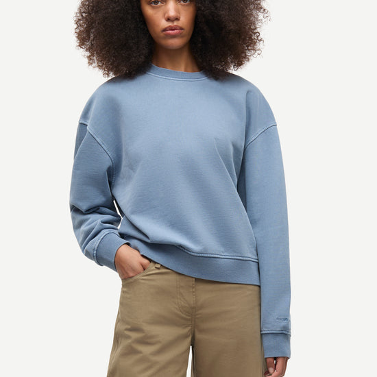 A woman with voluminous dark curly hair wearing a dusty blue crewneck sweatshirt and beige wide-leg pants, standing against a light gray studio background.