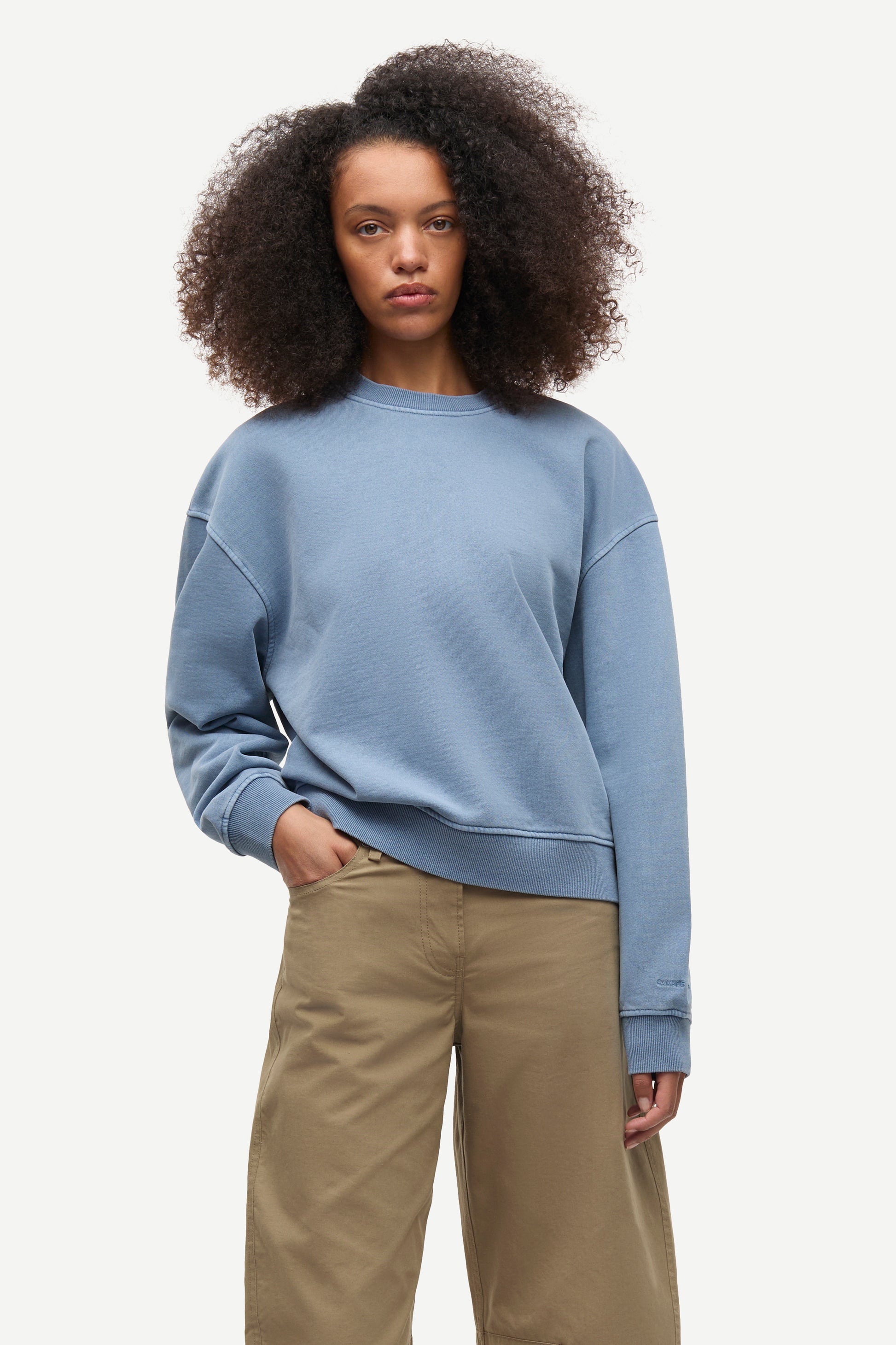 A woman with voluminous dark curly hair wearing a dusty blue crewneck sweatshirt and beige wide-leg pants, standing against a light gray studio background.
