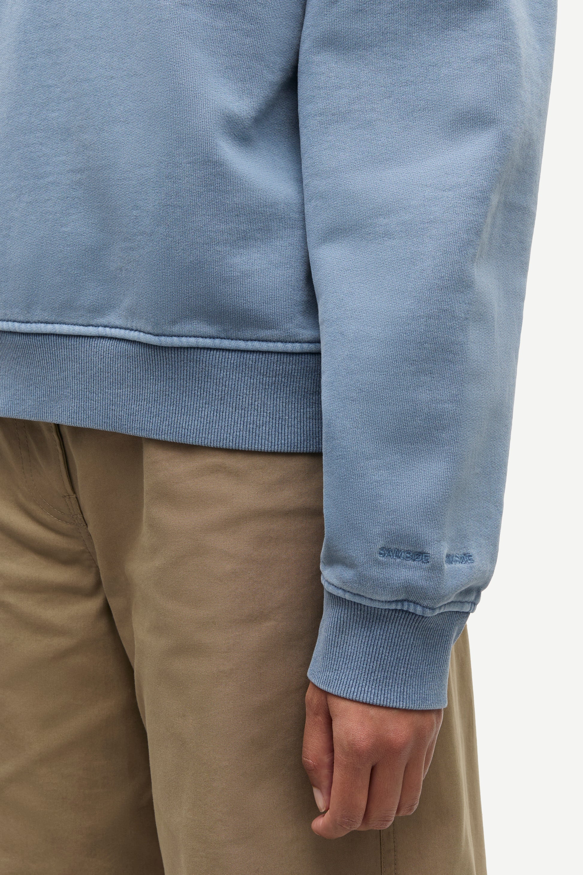 Close-up of a person wearing a light blue sweatshirt and tan pants, showing the ribbed cuffs and hem of the sweatshirt.