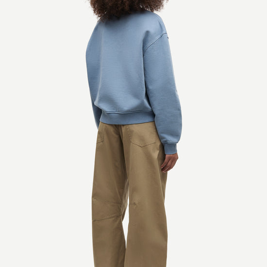 A child model shown from the back wearing a dusty blue sweatshirt and wide-leg khaki pants against a light gray studio background.
