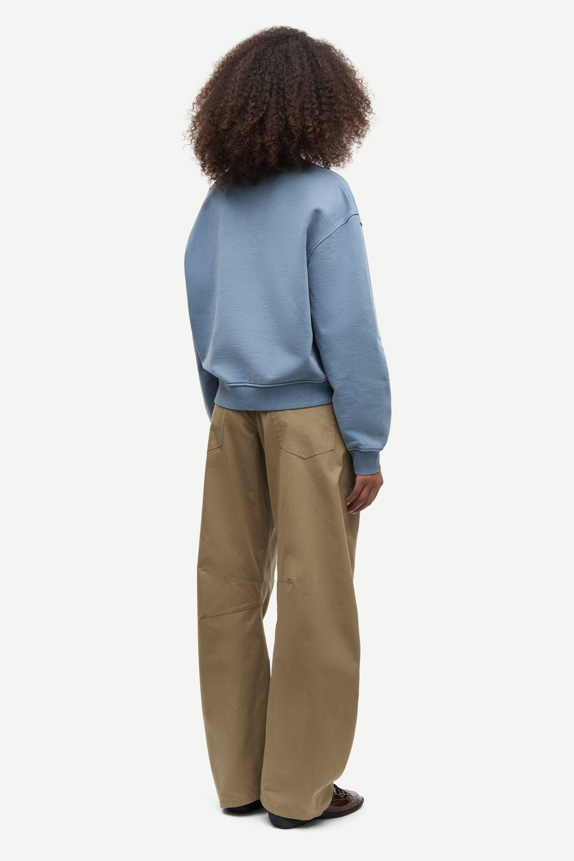 A child model shown from the back wearing a dusty blue sweatshirt and wide-leg khaki pants against a light gray studio background.