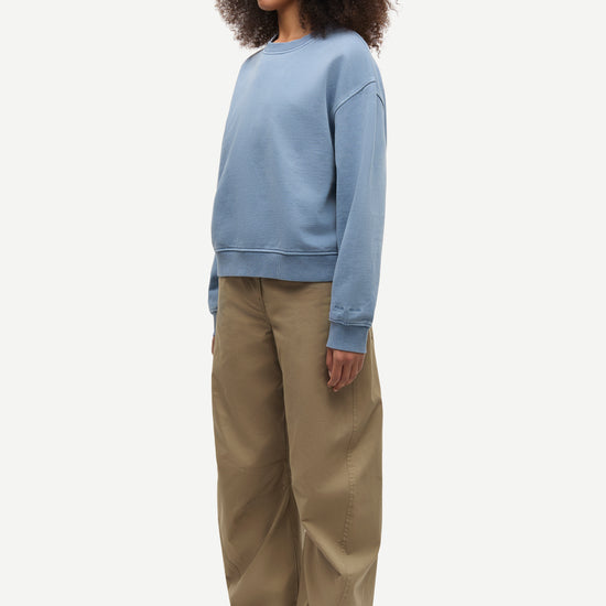 A model wearing a light blue crewneck sweatshirt, wide-leg khaki pants, and brown shoes, standing in profile against a plain light gray background.