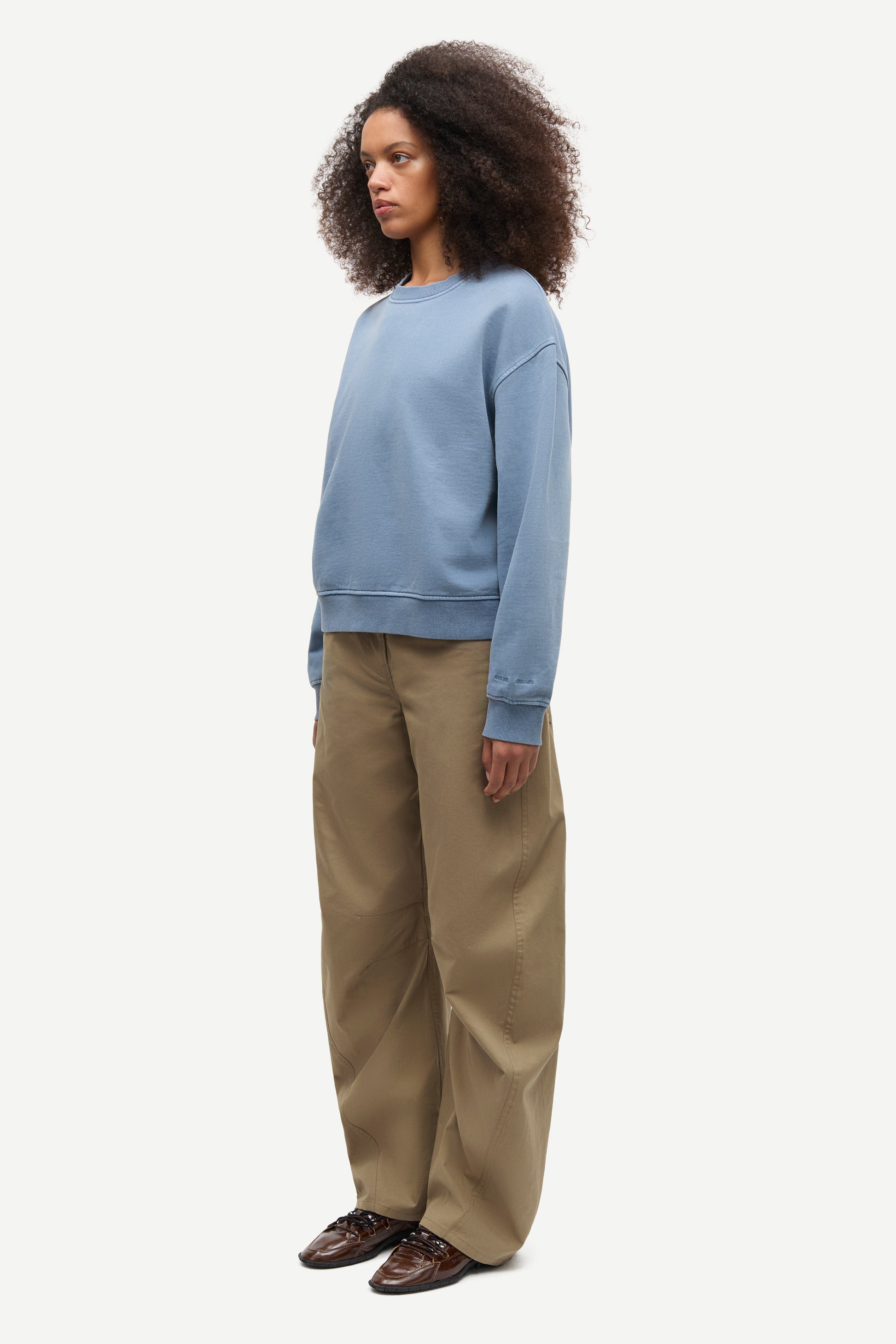 A model wearing a light blue crewneck sweatshirt, wide-leg khaki pants, and brown shoes, standing in profile against a plain light gray background.