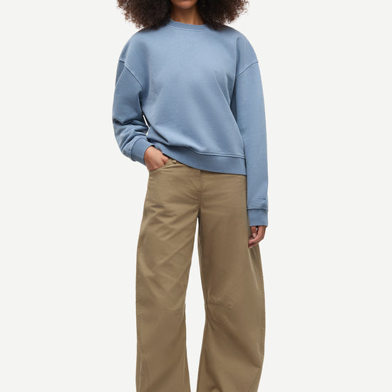 A model with curly dark hair stands against a light gray studio background wearing a light blue crewneck sweatshirt and wide-leg khaki pants.
