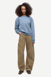 A model with curly dark hair stands against a light gray studio background wearing a light blue crewneck sweatshirt and wide-leg khaki pants.