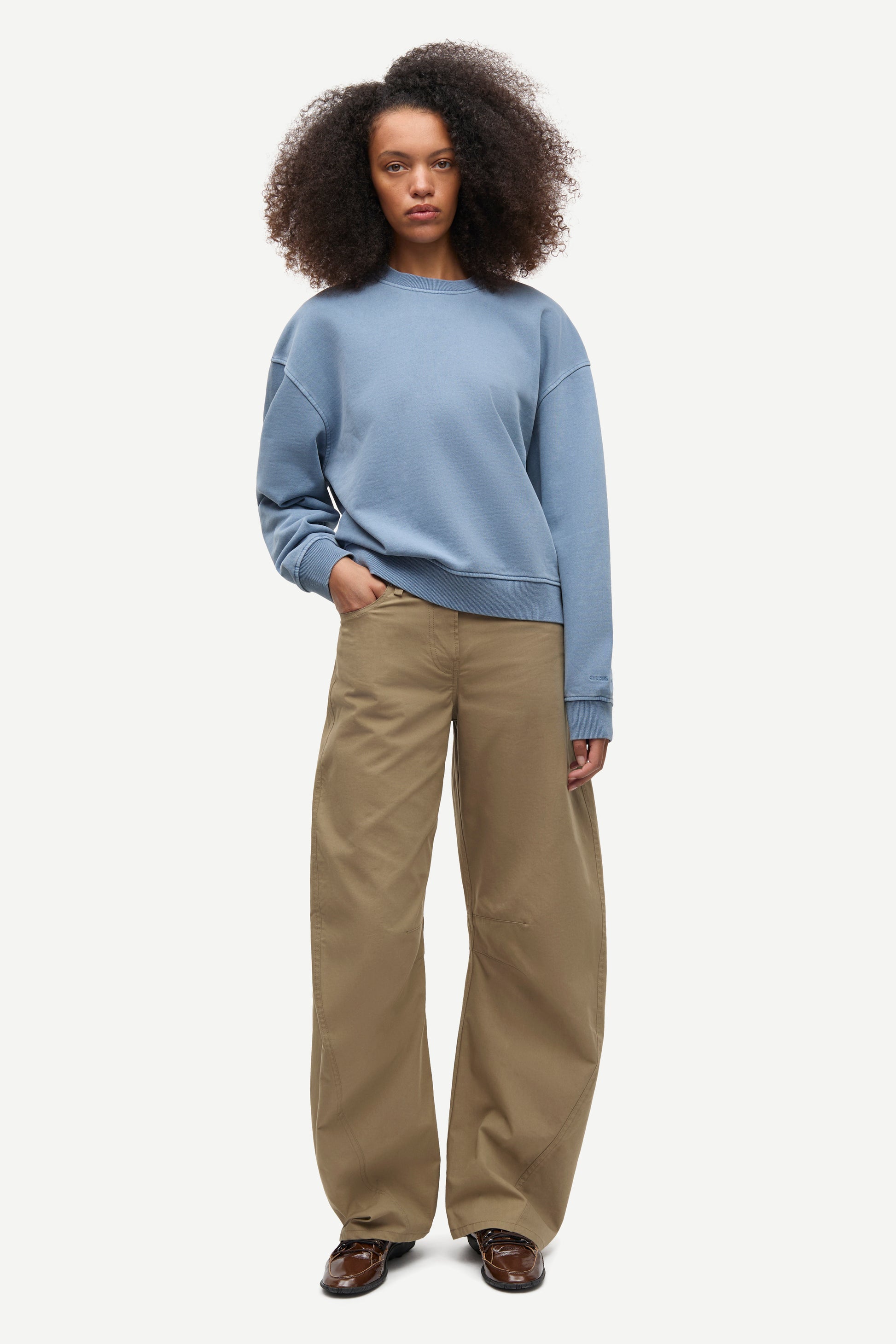 A model with curly dark hair stands against a light gray studio background wearing a light blue crewneck sweatshirt and wide-leg khaki pants.