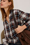 A woman wearing a long-sleeve plaid button-up shirt in muted blue, brown, and white tones, carrying a dark brown shoulder bag.
