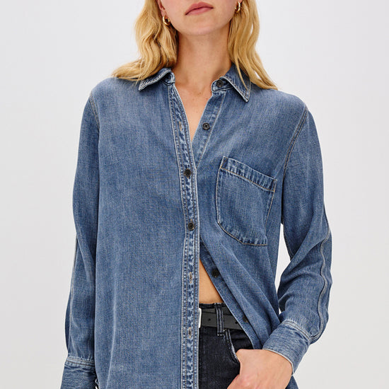 A woman wearing a medium-wash blue denim button-up shirt with a chest pocket, paired with dark jeans, against a plain light gray studio background.
