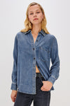 A woman wearing a medium-wash blue denim button-up shirt with a chest pocket, paired with dark jeans, against a plain light gray studio background.