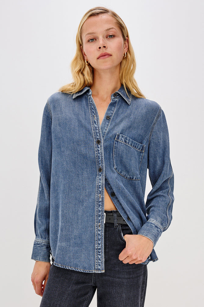 A woman wearing a medium-wash blue denim button-up shirt with a chest pocket, paired with dark jeans, against a plain light gray studio background.