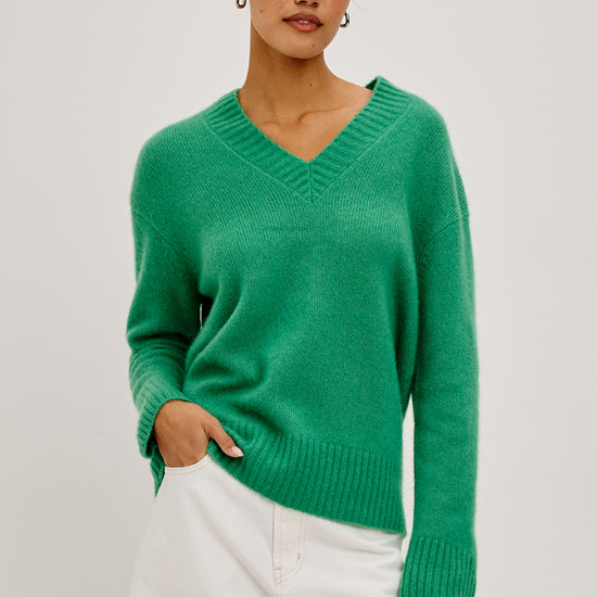 A woman modeling a bright green V-neck knit sweater with dropped shoulders, paired with white trousers, against a plain light gray studio background.