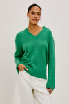 A woman modeling a bright green V-neck knit sweater with dropped shoulders, paired with white trousers, against a plain light gray studio background.
