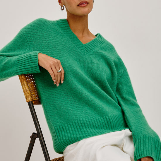 A woman wearing an oversized bright green V-neck knit sweater and white shorts, seated on a woven chair against a plain light gray studio background.
